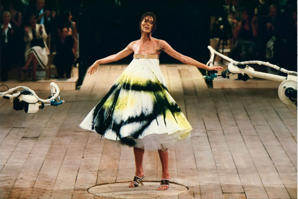 Alexander McQueen Spring/Summer 1999, collection No. 13. Shalom Harlow stands on a rotating platform as two industrial robot arms spray paint her white dress in black and yellow. Photographed by Robert Fairer. The show remains one of the most iconic moments in fashion history and a document of what Alexander McQueen stood for before the era of scalable luxury. | Photo Robert Fairer in Courtesy of Alexander McQueen
