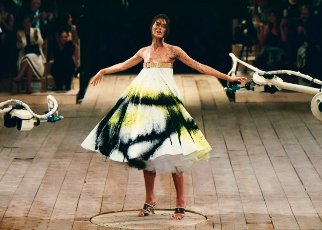 Alexander McQueen Spring/Summer 1999, collection No. 13. Shalom Harlow stands on a rotating platform as two industrial robot arms spray paint her white dress in black and yellow. Photographed by Robert Fairer. The show remains one of the most iconic moments in fashion history and a document of what Alexander McQueen stood for before the era of scalable luxury. | Photo Robert Fairer in Courtesy of Alexander McQueen