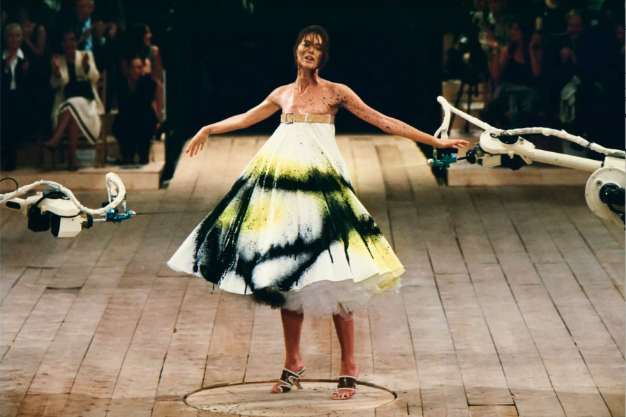 Alexander McQueen Spring/Summer 1999, collection No. 13. Shalom Harlow stands on a rotating platform as two industrial robot arms spray paint her white dress in black and yellow. Photographed by Robert Fairer. The show remains one of the most iconic moments in fashion history and a document of what Alexander McQueen stood for before the era of scalable luxury. | Photo Robert Fairer in Courtesy of Alexander McQueen