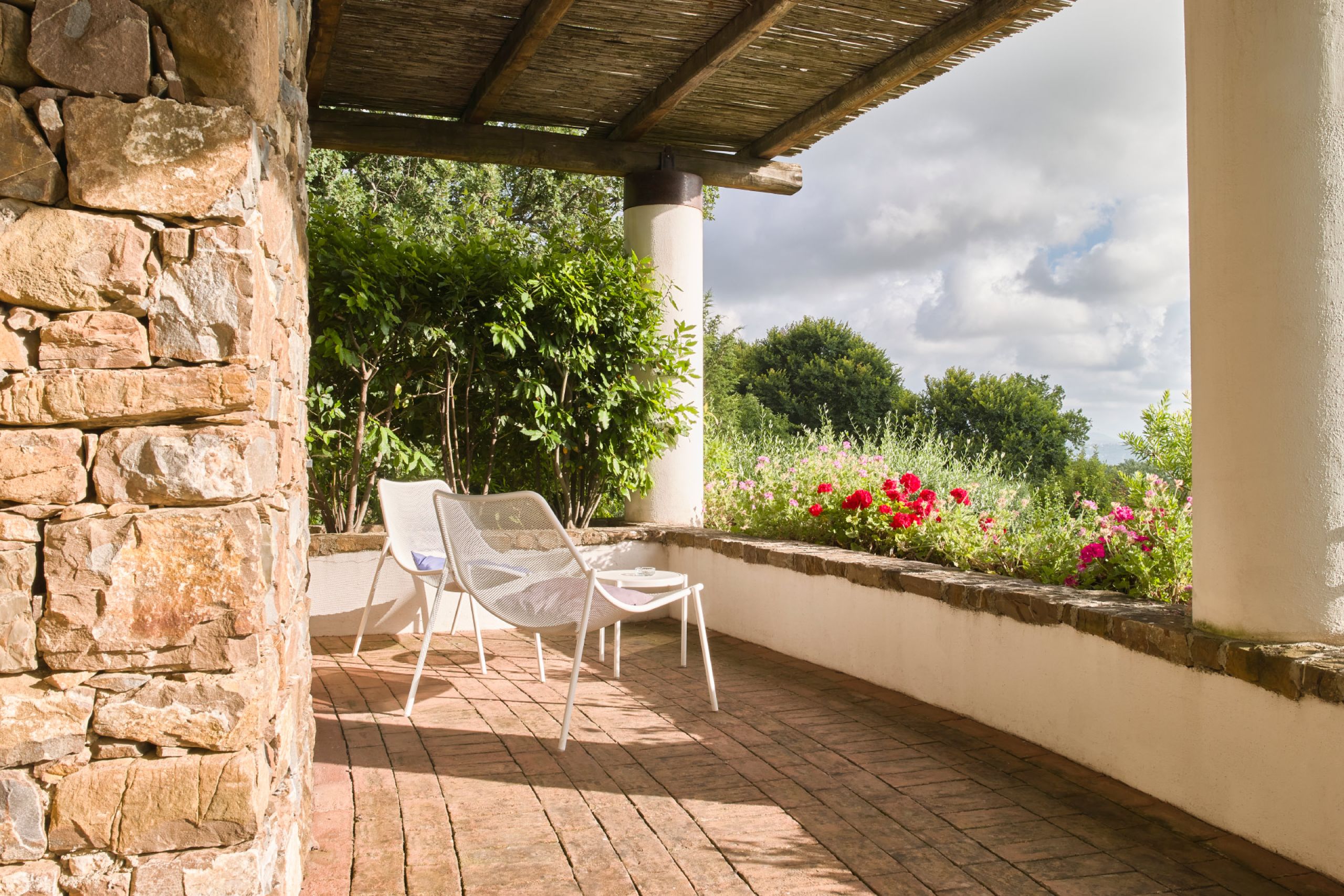 Private terrace of a suite at Borgo La Pietraia, local sandstone walls, reed pergola, white garden chairs and flowering Mediterranean garden, Cilento