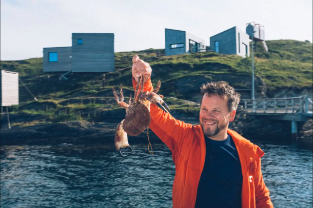 Harvard Lund holding a freshly caught crab in front of the monofunctional wooden cabins of The Arctic Hideaway on the island of Sørvær, Norway