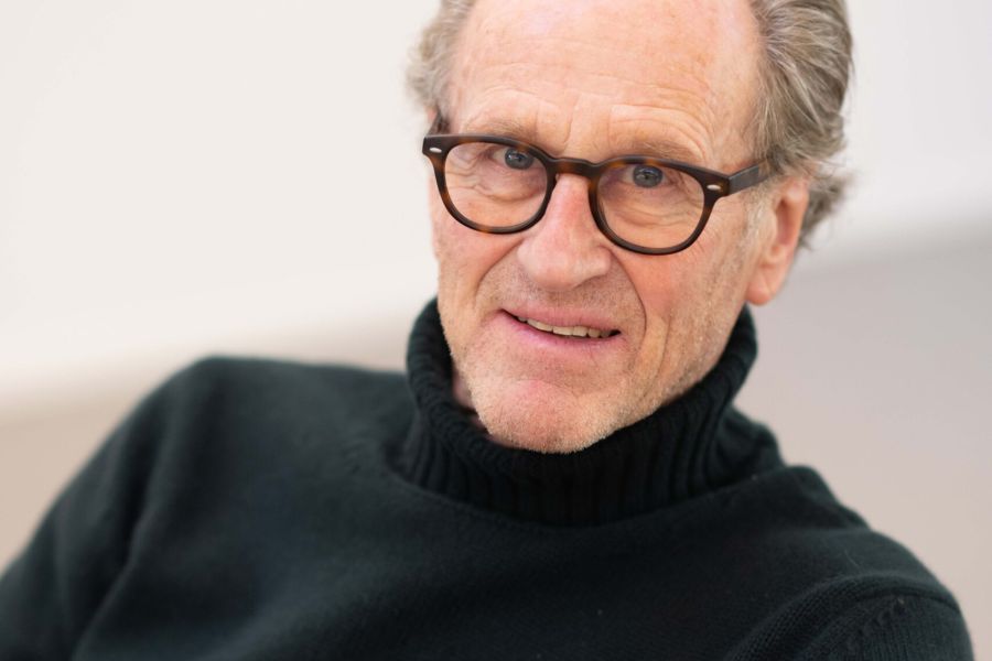A professional portrait of architect Matteo Thun, wearing a black turtleneck and tortoiseshell glasses, looking into the camera with a calm and focused expression. The image captures the essence of his "Eco, not Ego" philosophy and his commitment to botanical architecture.