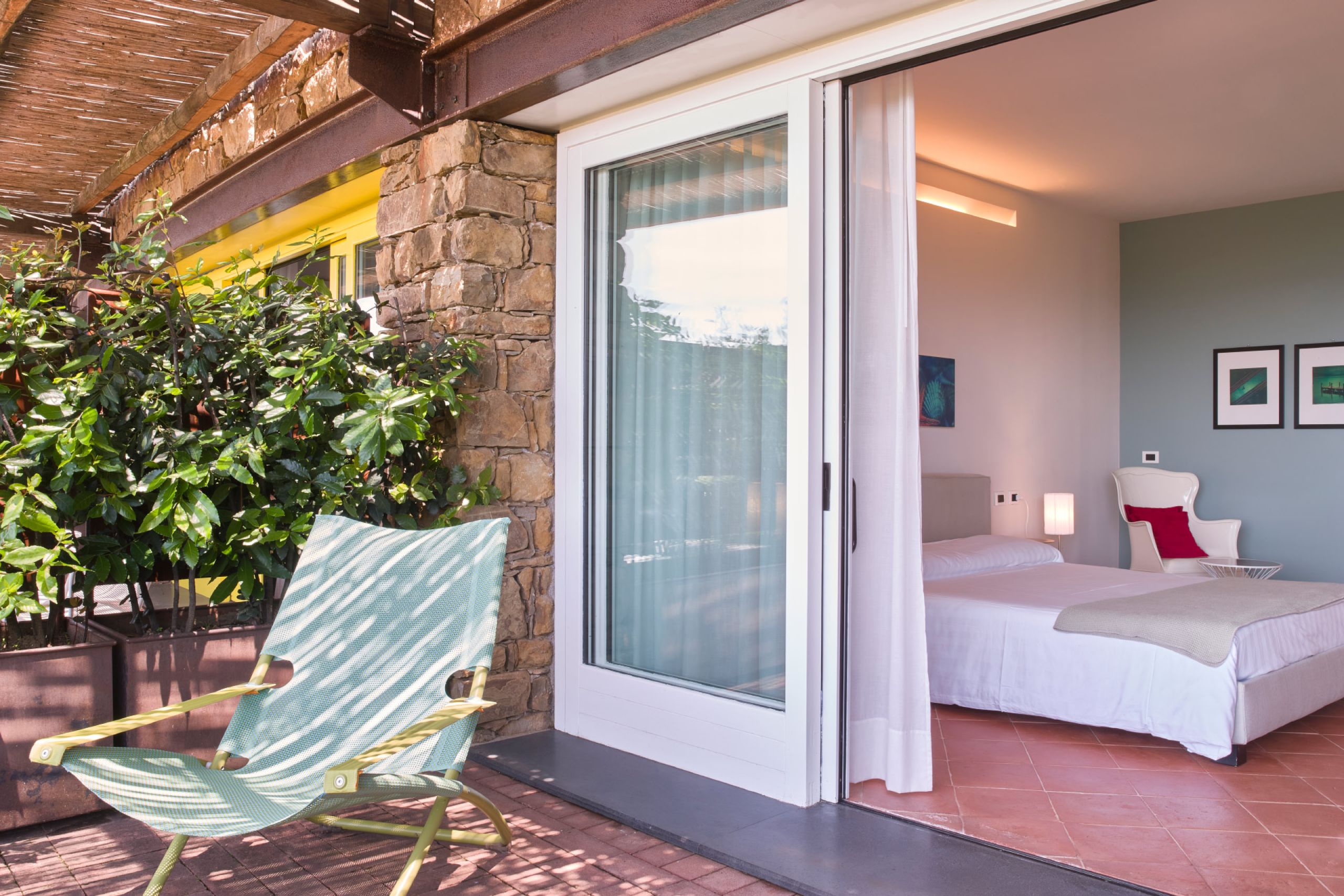 Junior Suite at Borgo La Pietraia, private terrace with green garden chair opening to light-filled room with local sandstone wall, yellow window accent and contemporary art, Cilento