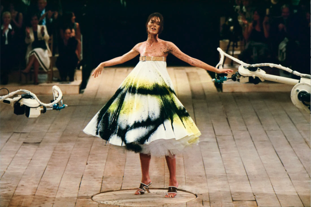 Alexander McQueen Spring/Summer 1999, collection No. 13. Shalom Harlow stands on a rotating platform as two industrial robot arms spray paint her white dress in black and yellow. Photographed by Robert Fairer. The show remains one of the most iconic moments in fashion history and a document of what Alexander McQueen stood for before the era of scalable luxury. | Photo Robert Fairer in Courtesy of Alexander McQueen