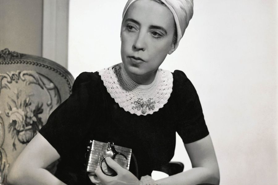 Elsa Schiaparelli wearing a black silk dress with crocheted collar of her own design and a white turban, seated in an upholstered chair holding a silver clutch. Vogue, 1940. Photograph by Fredrich Baker © Condé Nast via Getty Images.
