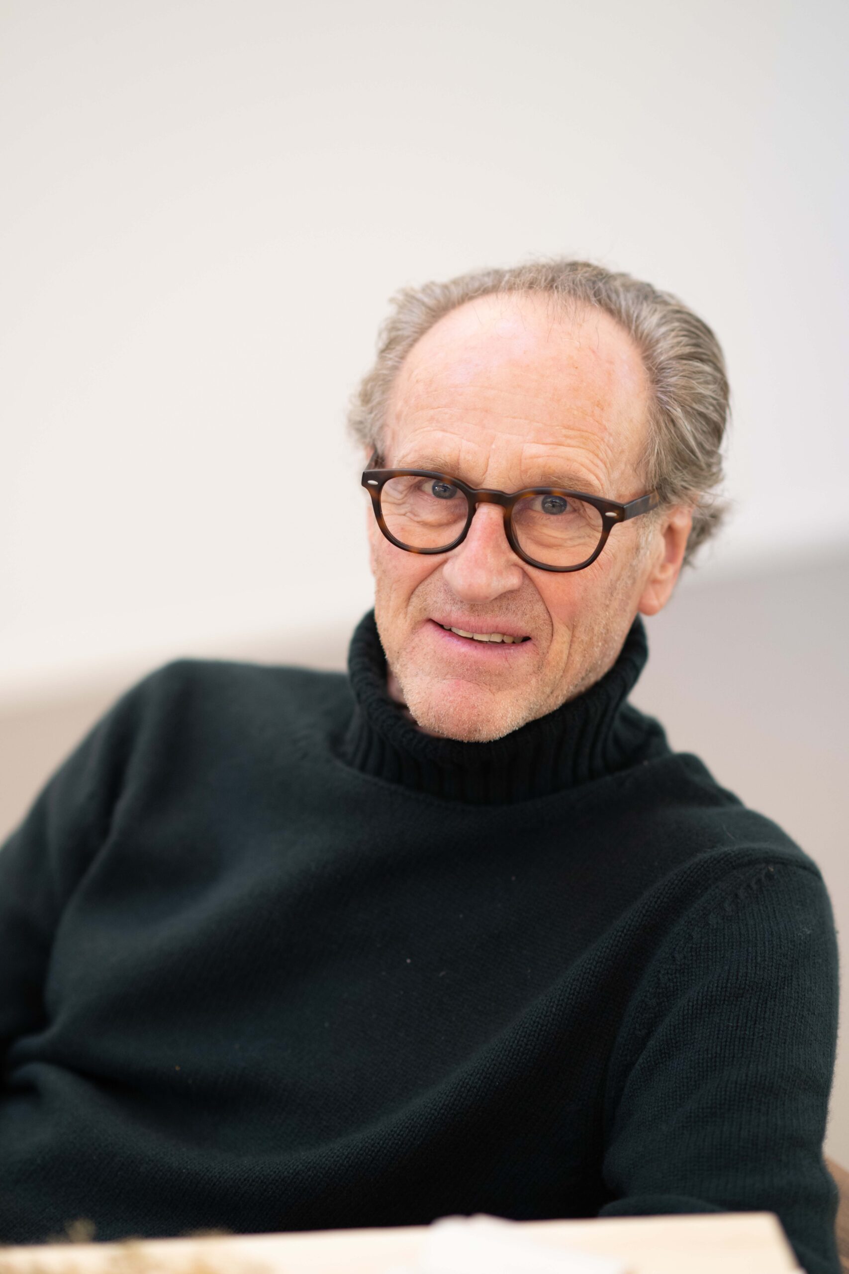 A professional portrait of architect Matteo Thun, wearing a black turtleneck and tortoiseshell glasses, looking into the camera with a calm and focused expression. The image captures the essence of his "Eco, not Ego" philosophy and his commitment to botanical architecture.