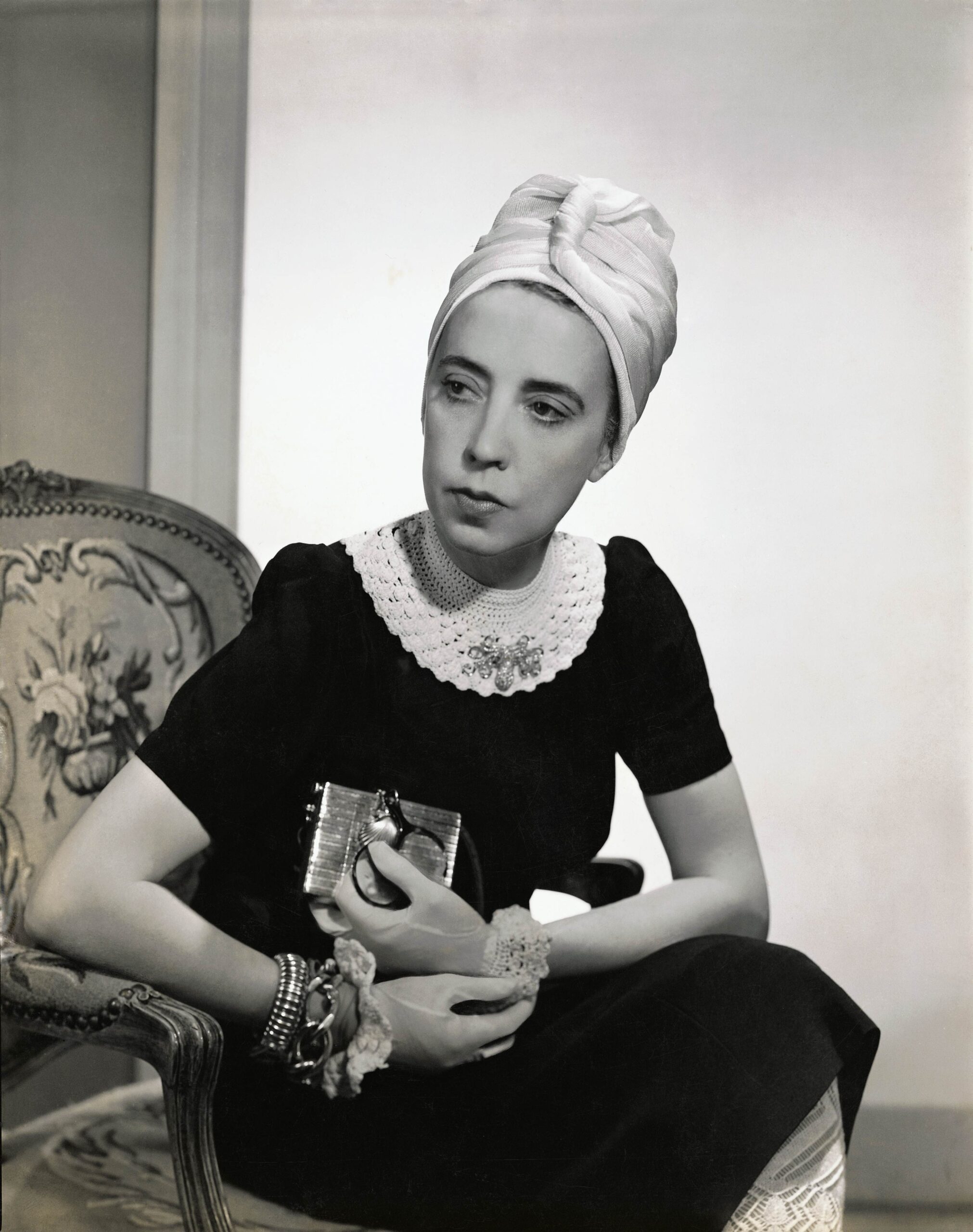 Elsa Schiaparelli wearing a black silk dress with crocheted collar of her own design and a white turban, seated in an upholstered chair holding a silver clutch. Vogue, 1940. Photograph by Fredrich Baker © Condé Nast via Getty Images.