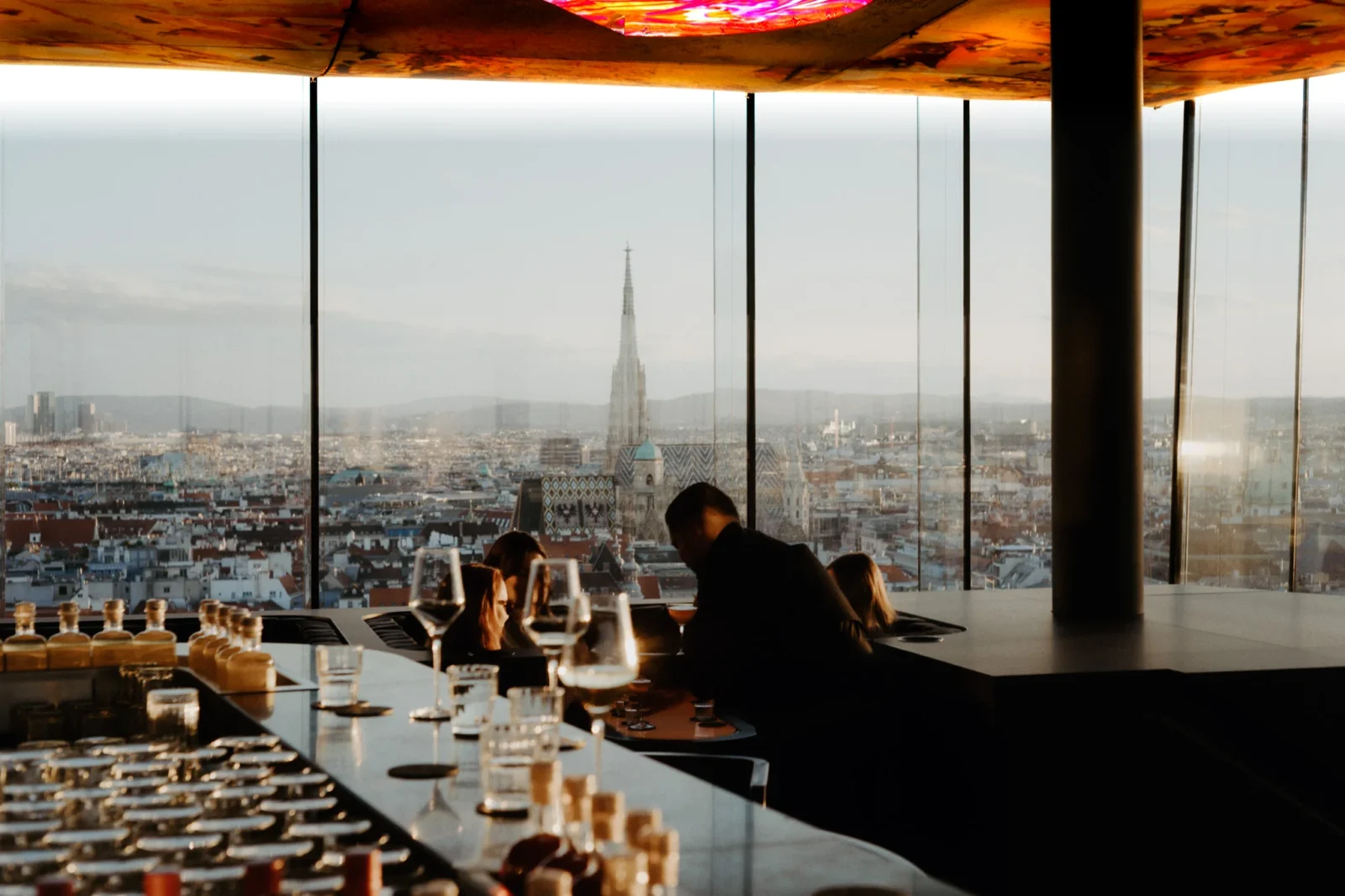 Guests at the bar of Das LOFT in the evening light beneath Pipilotti Rist's illuminated ceiling, with St Stephen's Cathedral rising over Vienna beyond the glass.