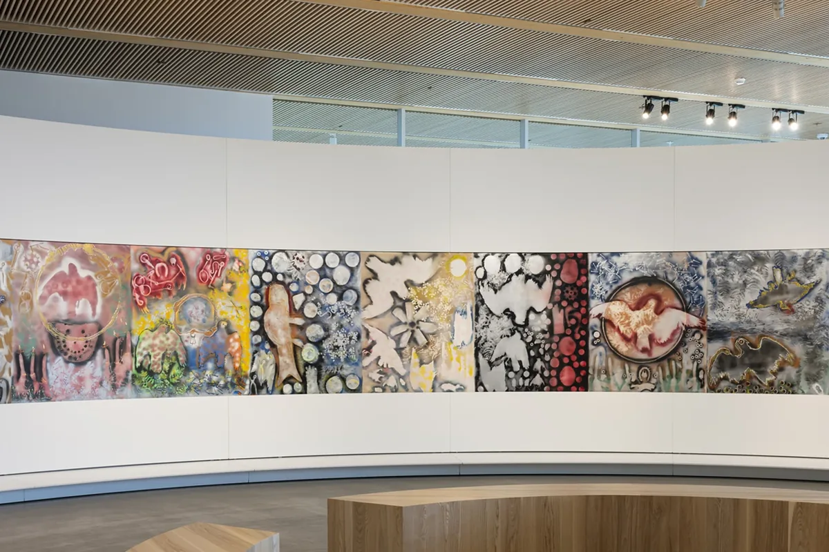 Jean Paul Riopelle's Tribute to Rosa Luxemburg (1992), a long, colourful painting of bird and object silhouettes, runs along the curved white wall of the circular room at the Espace Riopelle in Quebec City. Curved wooden benches stand in the foreground beneath a slatted wooden ceiling with track lights.