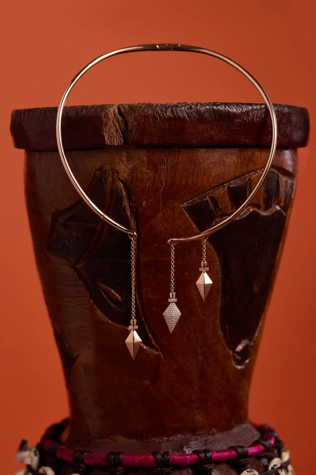 A Fullord Masai necklace in gold with spear-shaped drops, resting on a carved wooden vessel against a terracotta wall.