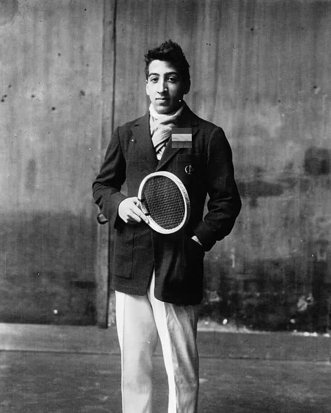 René Lacoste as a young tennis player in the 1920s, in a dark jacket and white trousers, holding a racket.