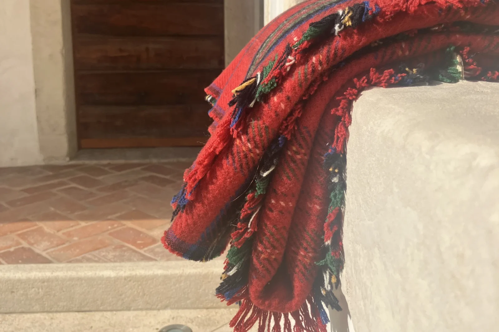 A red checked wool blanket with tufted, fringed edges lies folded over a stone wall in the courtyard of Lanificio Bottoli in Vittorio Veneto.