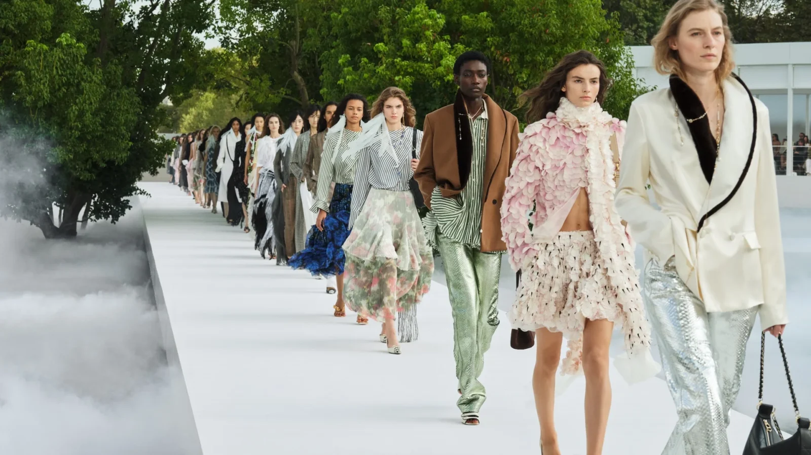 Models walking a white Christian Dior runway lined with trees, the first look in an ivory jacket and silver trousers