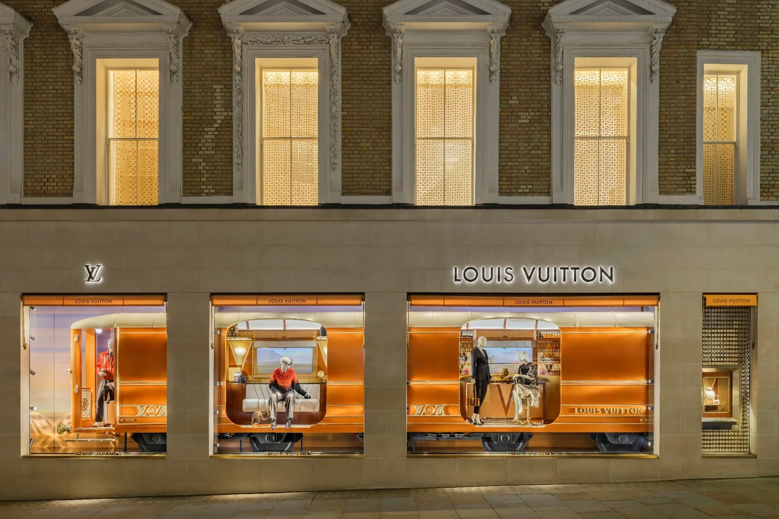 Evening view of a Louis Vuitton store, its three illuminated windows framed in copper below a brick façade