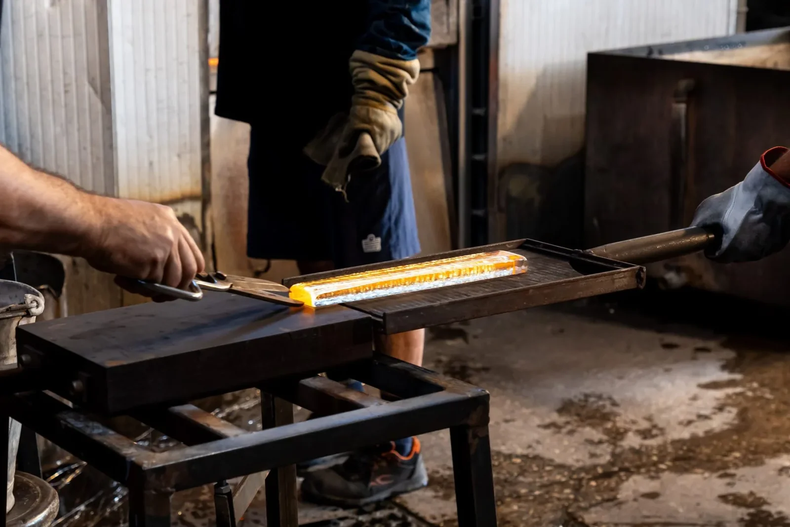 A glassmaker in heavy gloves draws a bar of glowing molten glass across a steel bench at the furnace.