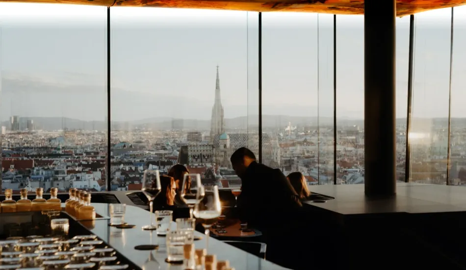 Guests at the bar of Das LOFT in the evening light beneath Pipilotti Rist's illuminated ceiling, with St Stephen's Cathedral rising over Vienna beyond the glass.