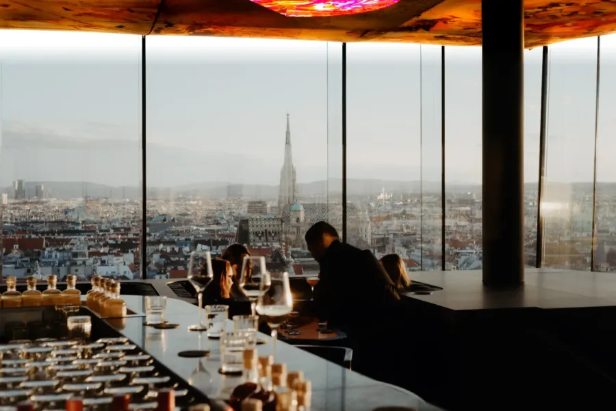 Guests at the bar of Das LOFT in the evening light beneath Pipilotti Rist's illuminated ceiling, with St Stephen's Cathedral rising over Vienna beyond the glass.